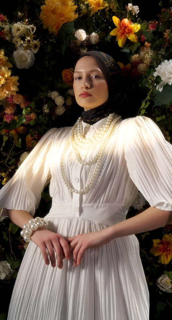 "Çiçeklerle bezenmiş fon önünde inci detaylı beyaz elbise ve siyah desenli şal ile poz veren zarif kadın model"