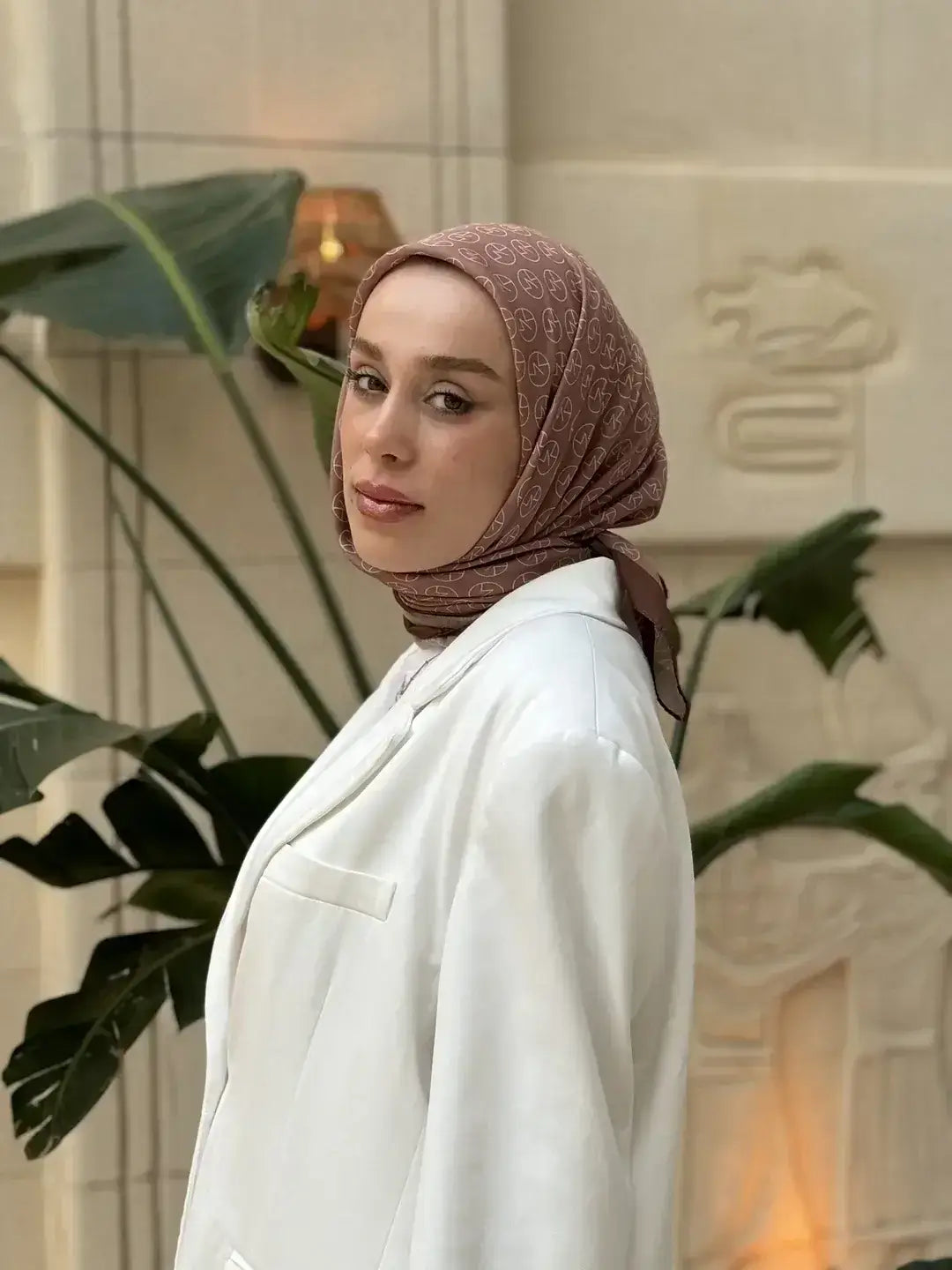 MK logolu kahverengi saten eşarp takan kadın model, beyaz blazer ceketle modern tesettür stili sergiliyor
