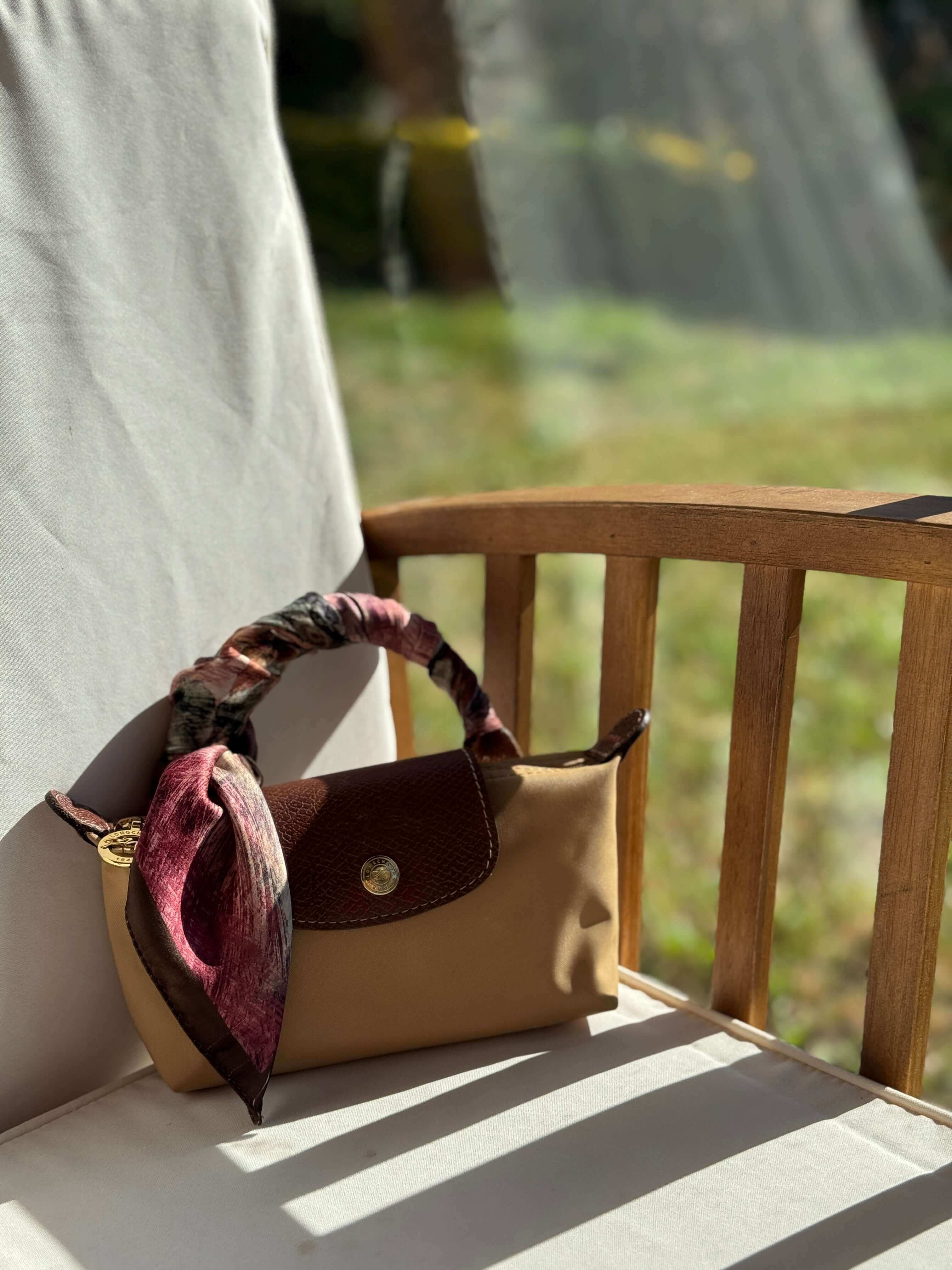 "Doğal ışıkta ahşap sandalyeye yerleştirilmiş çantaya zarifçe bağlanmış bordo tonlarında desenli fular – stilinizi tamamlayan şık ve özgün bir aksesuar."