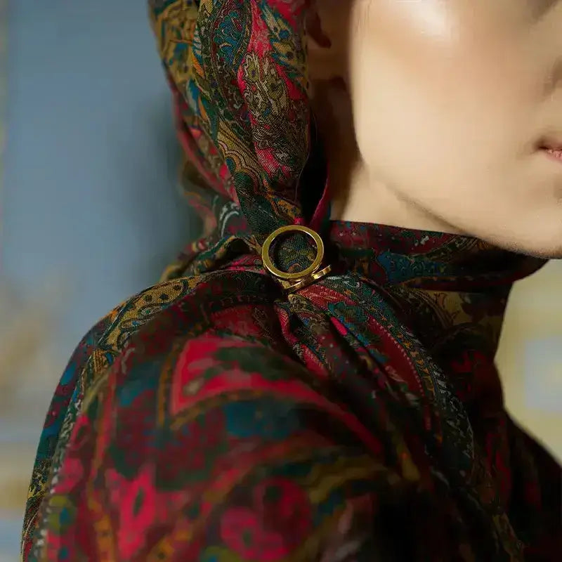 “Moda Nûda etnik desenli şal – altın renkli şal yüzüğüyle sabitlenmiş, kırmızı ve zümrüt tonlarında klasik detaylı ipek eşarp görünümü”