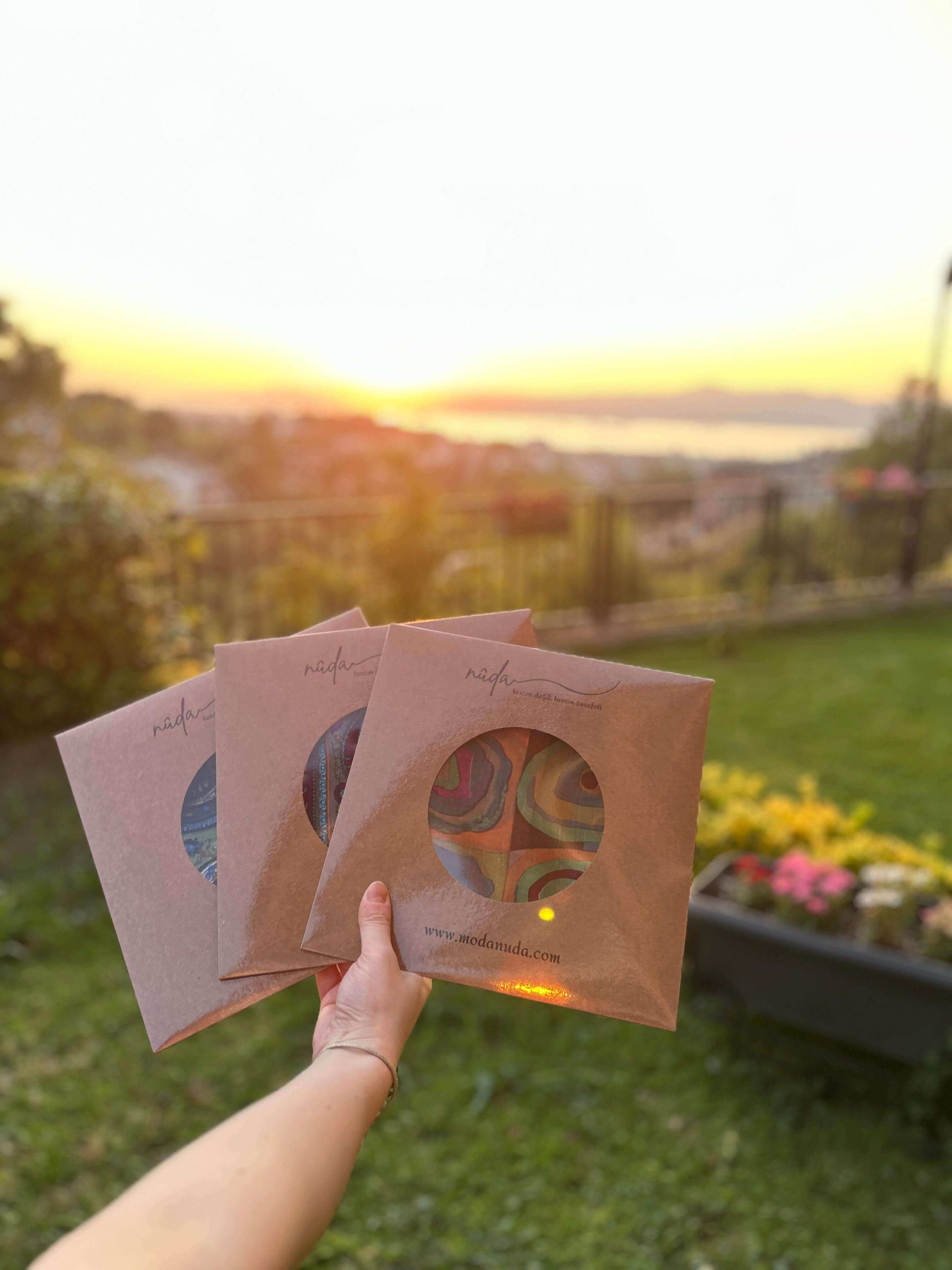 “Moda Nûda fular setleri – çim alanda gün batımında elde tutulan renkli desenli üç adet fular paketi”