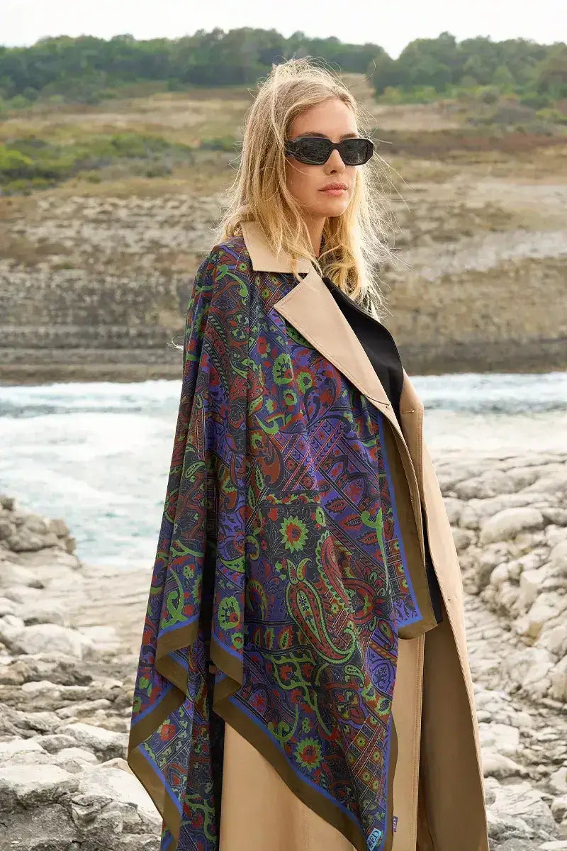 “Moda Nûda etnik desenli şal – mavi, yeşil ve kırmızı motifli büyük ebat şal, doğa manzarasında modern trençkotla kombinlenmiş”